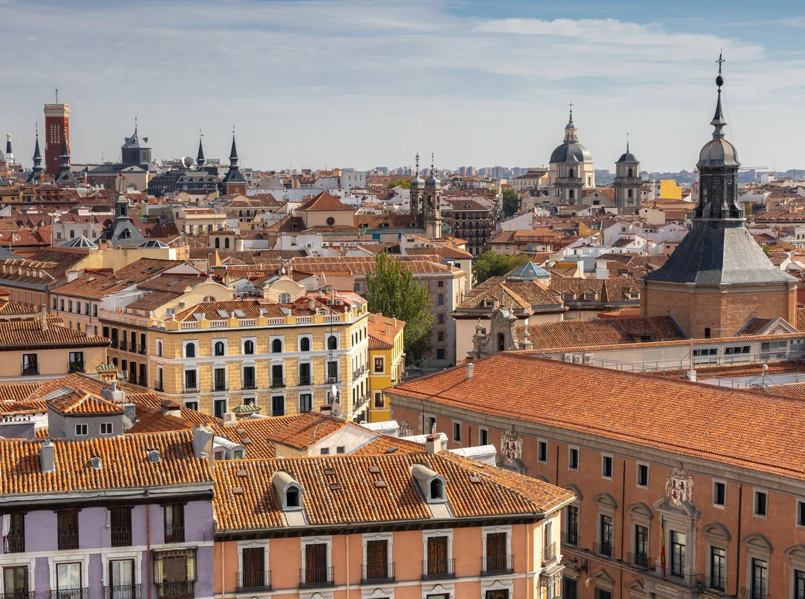 Terracotta-tiled rooftops, church towers and pastel facades across the historic centre of Madrid under a pale sky.